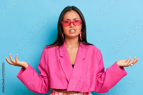 Indoor shot of hesitant clueless brunette woman shrug shoulders spread palms aside has doubt while make decision stands puzzled wears modern pink jacket and transparent spectacles isolated over blue
