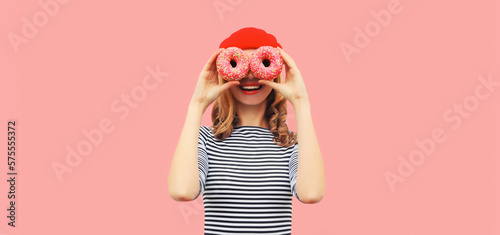 Portrait of happy funny smiling woman covering her eyes looking for something looking through donuts as binoculars having fun on pink background
