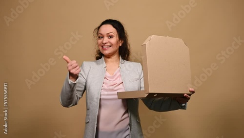 Beautiful happy woman opens a cardboard flat box with hot Italian pizza, shows thumb up and tasty gesture, kisses fingers and says bellissimo, isolated on beige background. Gourmet culinary concept photo