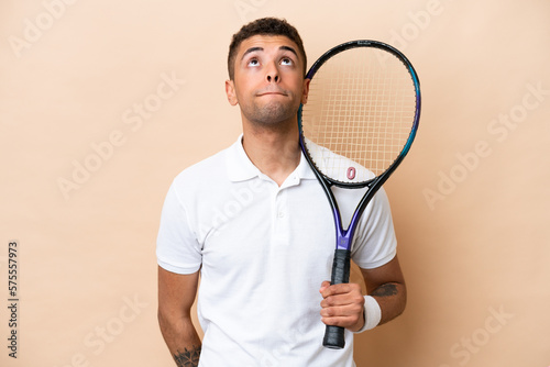 Young brazilian handsome man playing tennis isolated on beige background and looking up © luismolinero