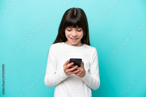 Little caucasian girl isolated on blue background sending a message with the mobile