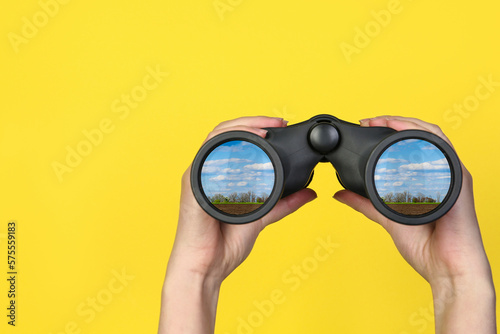 Fotografie Woman holding binoculars on yellow background, closeup