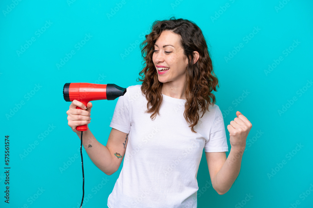 Young caucasian woman holding a hairdryer isolated on blue background celebrating a victory Young caucasian woman holding a hairdryer isolated on blue background celebrating a victory