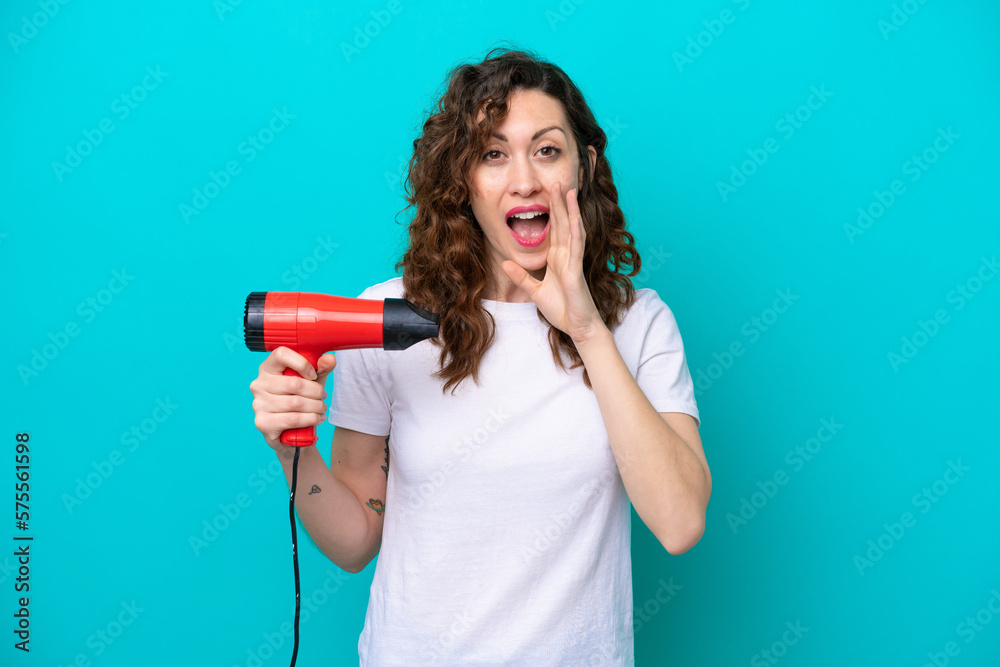 Young caucasian woman holding a hairdryer isolated on blue background shouting with mouth wide open Young caucasian woman holding a hairdryer isolated on blue background shouting with mouth wide open