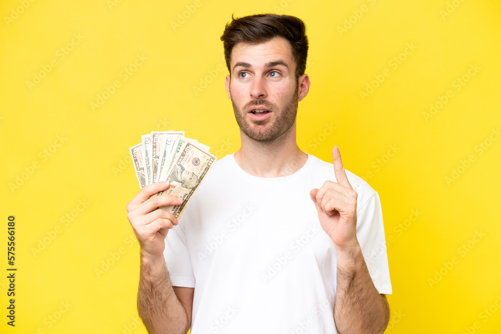 Young caucasian man taking a lot of money isolated on yellow background thinking an idea pointing the finger up Young caucasian man taking a lot of money isolated on yellow background thinking an idea pointing the finger up
