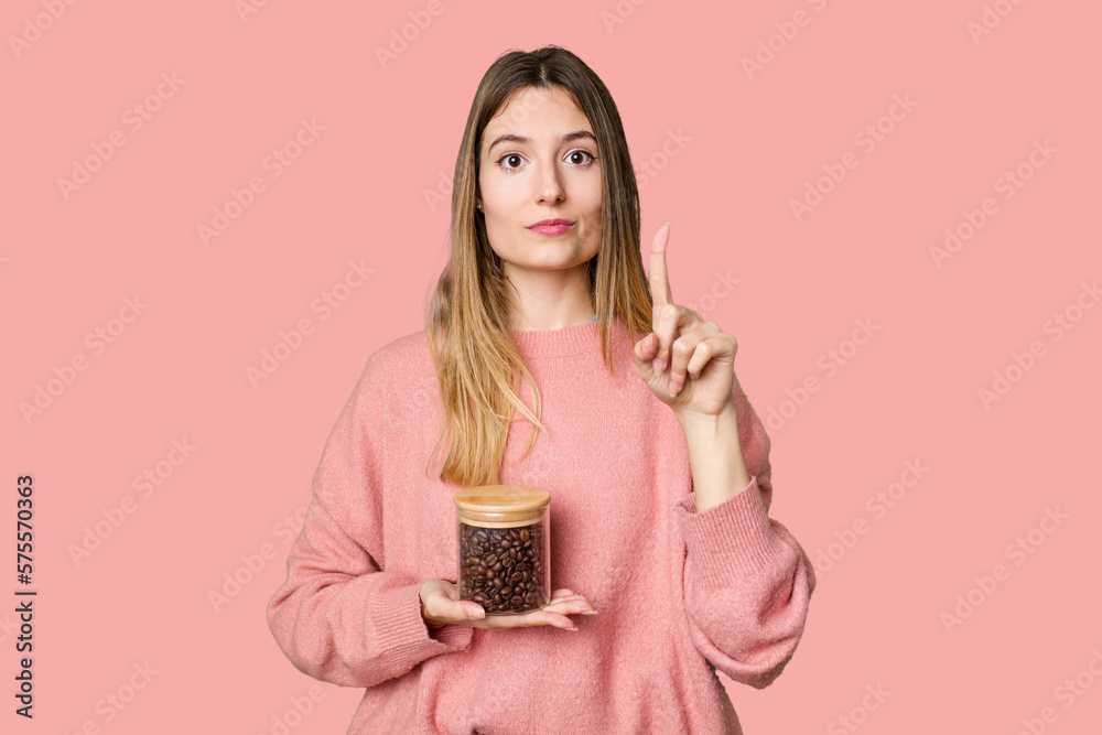 Young woman holding a container of coffee beans, symbolizing the energy and invigoration of this beloved beverage showing number one with finger. Young woman holding a container of coffee beans, symbolizing the energy and invigoration of this beloved beverage showing number one with finger.