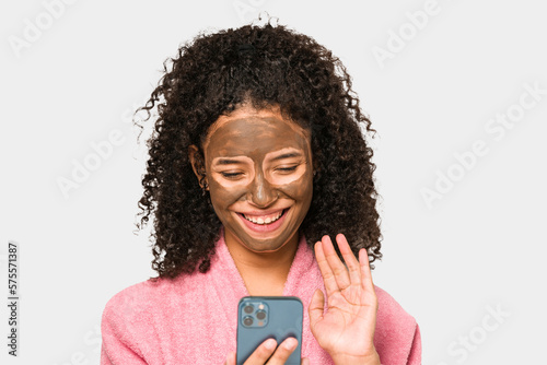 Young african american woman talking on phone while she is applying a facial mask isolated