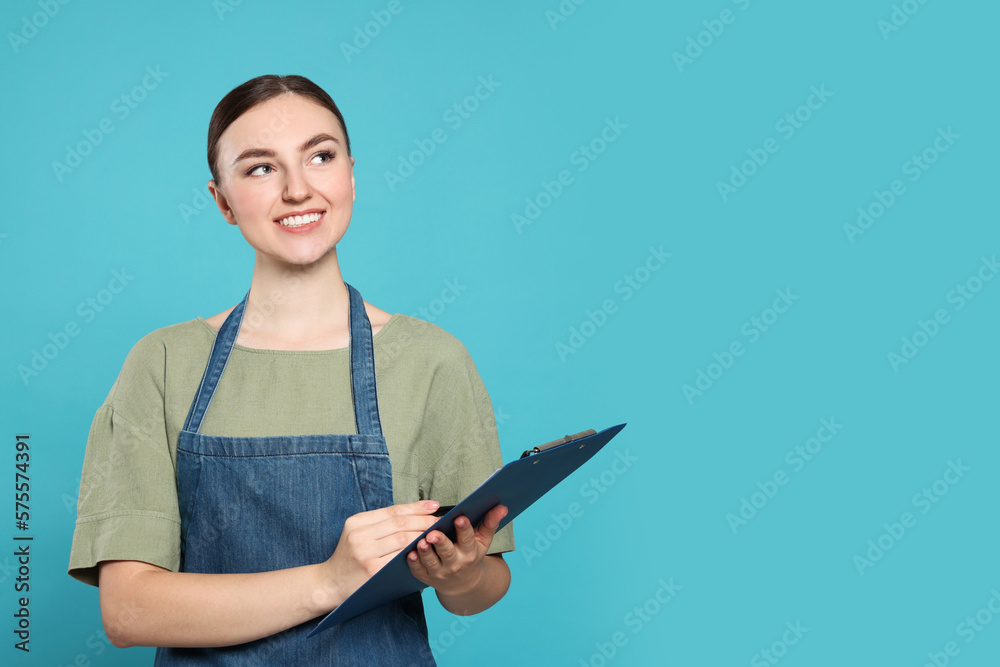 Beautiful young woman in clean denim apron with clipboard on light blue background. Space for text Beautiful young woman in clean denim apron with clipboard on light blue background. Space for text