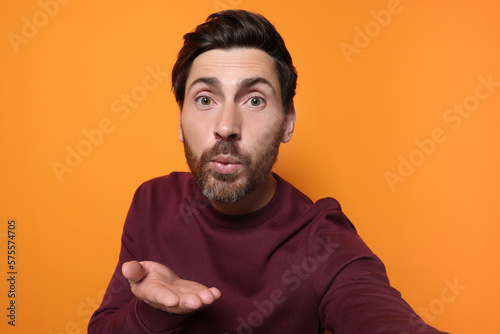Handsome man blowing kiss while taking selfie on orange background