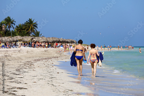 Fototapeta Naklejka Na Ścianę i Meble -  People walking by the sand on tropical beach on coconut palm trees background, two women in bikini in foreground. Ocean coast, tourist resort on Caribbean island