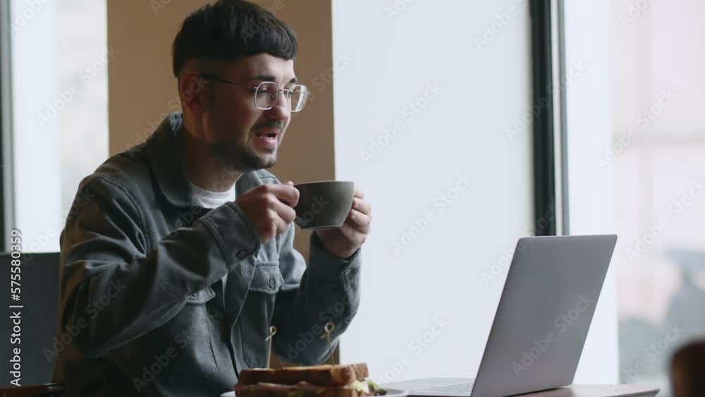Freelancer drinking coffee while working in cafe. Smiling young man in glasses using laptop in coffeehouse. Cheerful guy typing on computer keyboard in the restaurant Freelancer drinking coffee while working in cafe. Smiling young man in glasses using laptop in coffeehouse. Cheerful guy typing on computer keyboard in the restaurant