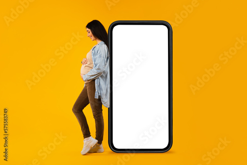 Happy pregnant woman leaning on huge blank cellphone and hugging her belly, advertising mobile app or website, mockup © Prostock-studio