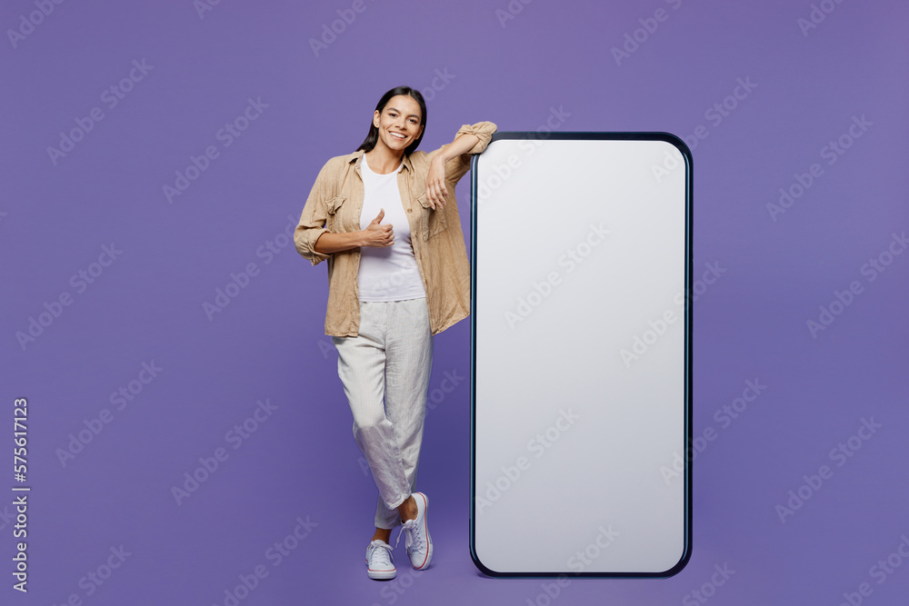 Full body smiling young latin woman wear light shirt casual clothes big huge blank screen mobile cell phone smartphone with area show thumb up isolated on plain pastel purple color background studio. Full body smiling young latin woman wear light shirt casual clothes big huge blank screen mobile cell phone smartphone with area show thumb up isolated on plain pastel purple color background studio.