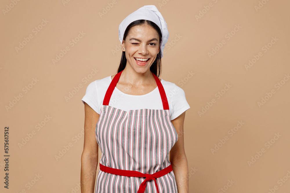 Young cheerful smiling happy housewife housekeeper chef cook baker latin woman wear striped apron toque hat looking camera wink isolated on plain pastel light beige background Cooking food concept. Young cheerful smiling happy housewife housekeeper chef cook baker latin woman wear striped apron toque hat looking camera wink isolated on plain pastel light beige background Cooking food concept.