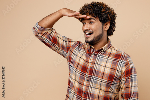 Side view optimist Indian man wears brown shirt casual clothes hold hand at forehead look far away distance isolated on plain pastel light beige background studio portrait. People lifestyle concept.