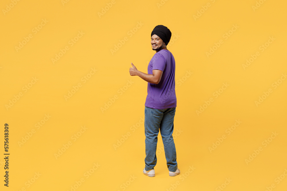 Full body back view happy smiling fun devotee Sikh Indian man ties his traditional turban dastar wear purple t-shirt showing thumb up like gesture isolated on plain yellow background studio portrait. Full body back view happy smiling fun devotee Sikh Indian man ties his traditional turban dastar wear purple t-shirt showing thumb up like gesture isolated on plain yellow background studio portrait.
