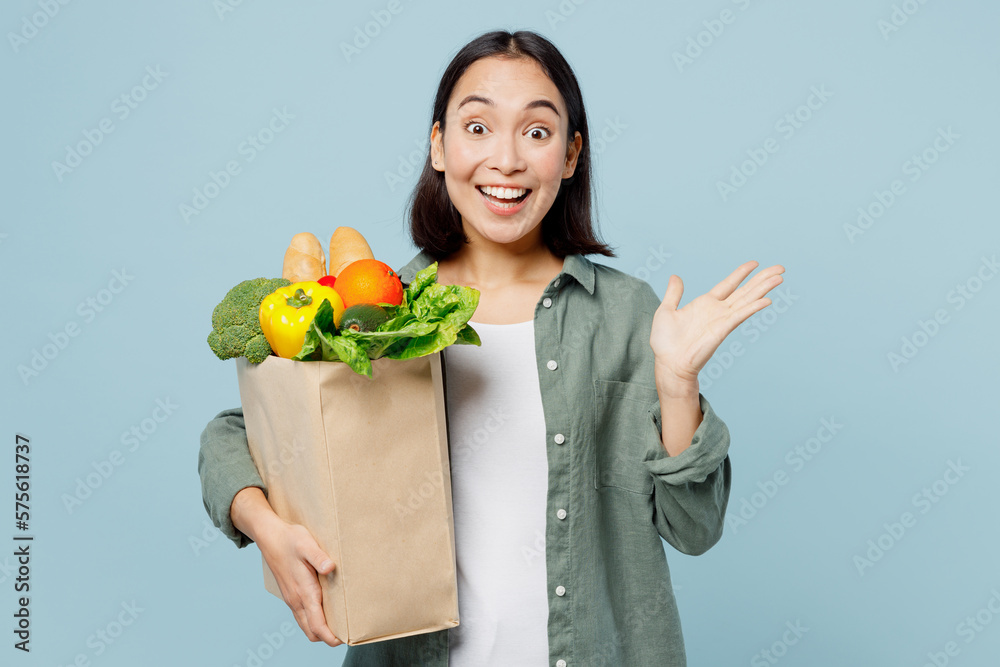 Young excited amazed fun woman wear casual clothes hold brown paper bag with food products spread hand isolated on plain blue cyan background studio portrait. Delivery service from shop or restaurant. Young excited amazed fun woman wear casual clothes hold brown paper bag with food products spread hand isolated on plain blue cyan background studio portrait. Delivery service from shop or restaurant.