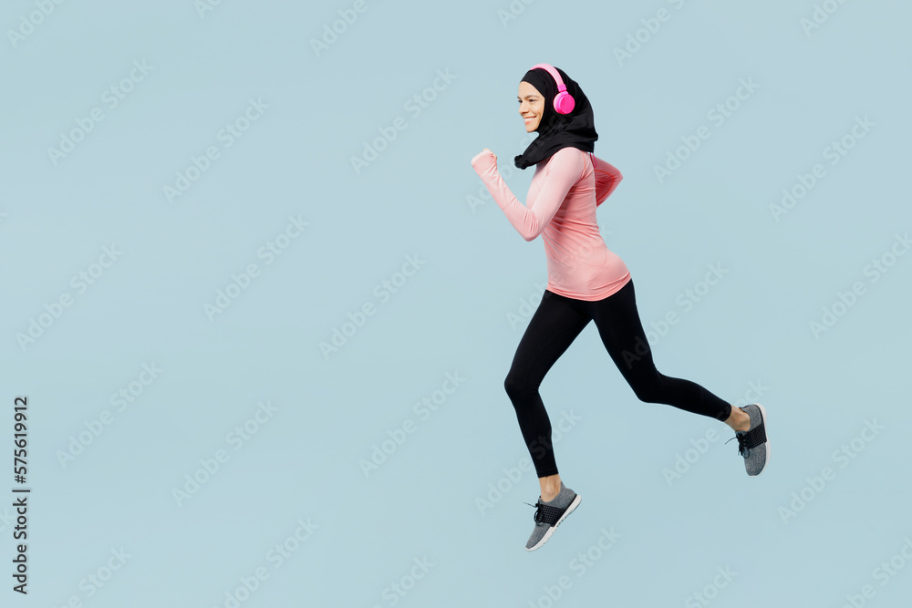 Full body young asian muslim fitness trainer sporty woman wear pink abaya hijab headphones listen music spend time in gym run jump high isolated on plain blue background studio. sport fit abs concept.
