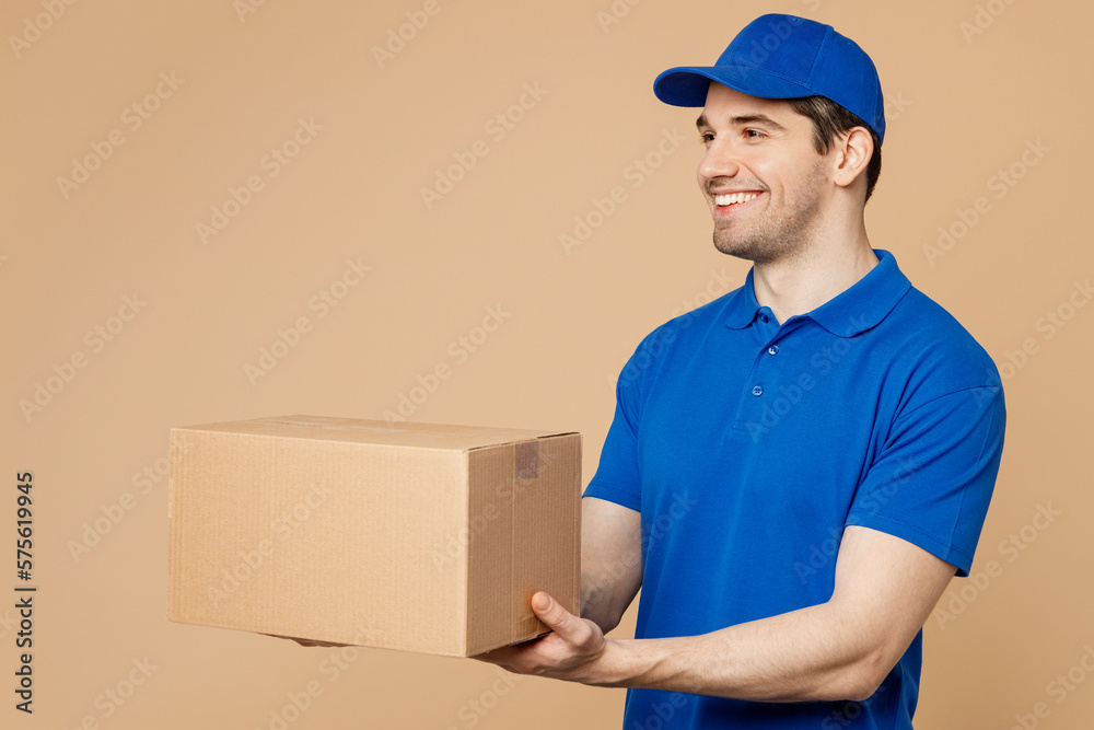 Side view professional happy delivery guy employee man wear blue cap t-shirt uniform workwear work as dealer courier hold give cardboard box isolated on plain light beige background. Service concept. Side view professional happy delivery guy employee man wear blue cap t-shirt uniform workwear work as dealer courier hold give cardboard box isolated on plain light beige background. Service concept.