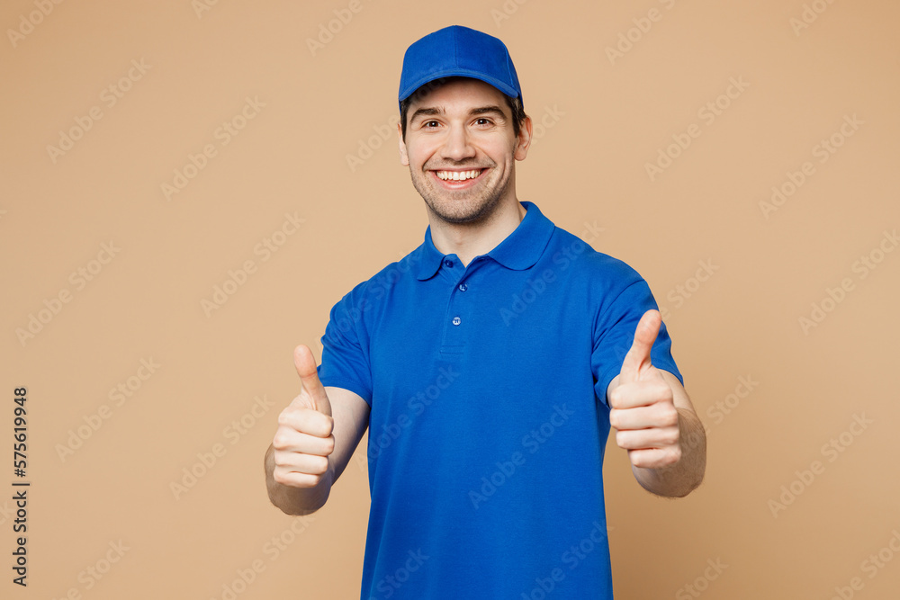 Professional satisfied delivery guy employee man wear blue cap t-shirt uniform workwear work as dealer courier showing thumb up like gesture isolated on plain light beige background. Service concept.