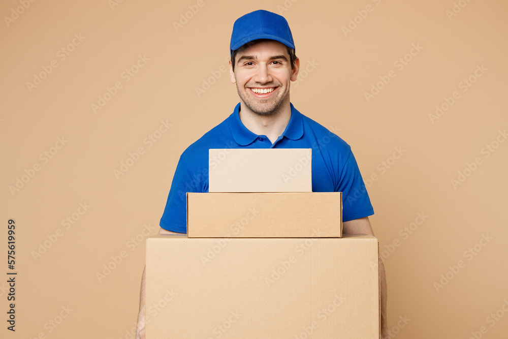 Professional happy delivery guy employee man wear blue cap t-shirt uniform workwear work as dealer courier hold stack cardboard blank boxes isolated on plain light beige background. Service concept. Professional happy delivery guy employee man wear blue cap t-shirt uniform workwear work as dealer courier hold stack cardboard blank boxes isolated on plain light beige background. Service concept.