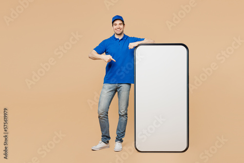 Full body happy delivery guy employee man wear blue cap t-shirt uniform workwear work as dealer courier point on big huge blank screen mobile cell phone with area isolated on plain beige background. photo
