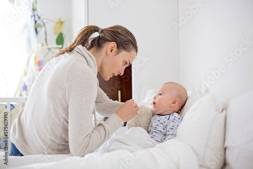 Cute sick child, baby boy, staying in bed, mom giving him medicine and checking on him
