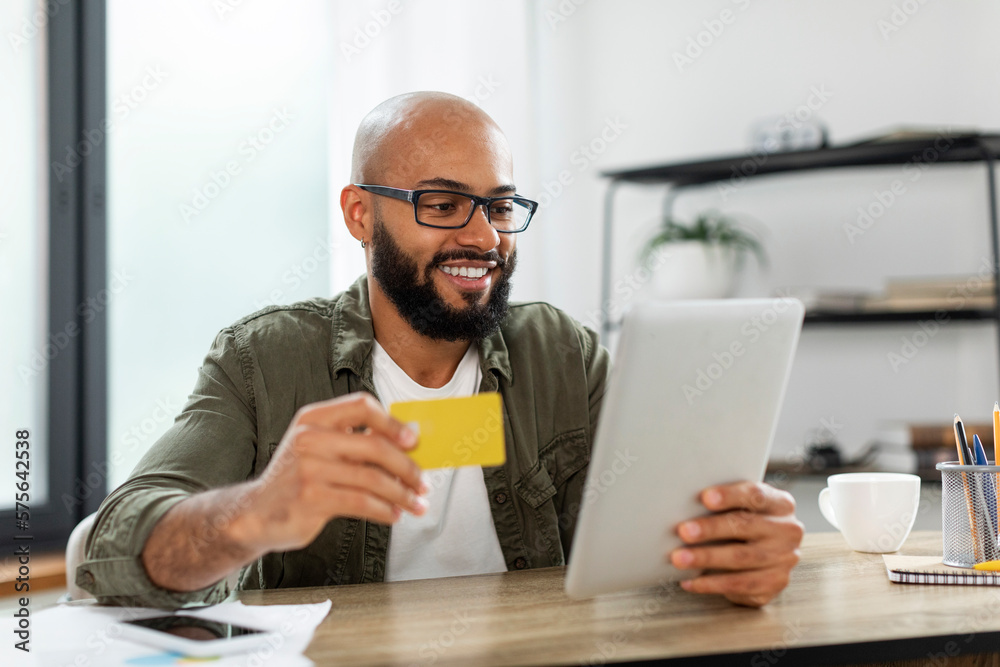 Online shopping. Excited latin businessman using tablet and credit card, making payment via bank app, sitting at desk Online shopping. Excited latin businessman using tablet and credit card, making payment via bank app, sitting at desk