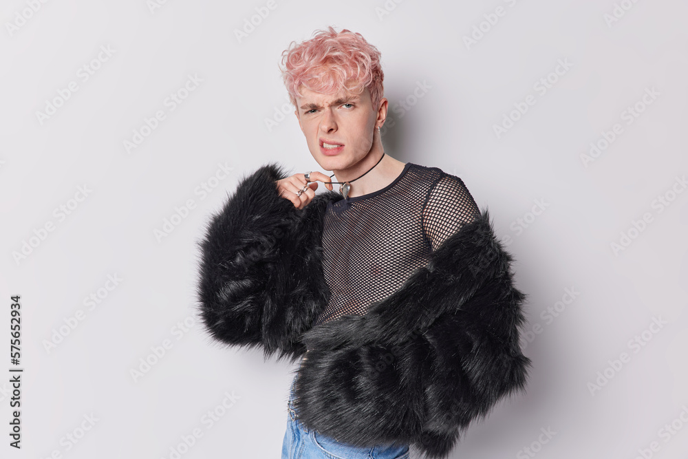Gay man in net shirt and black fur coat smirks face keeps hand on necklace posseses both masculine feminine characteristics changed his appearance poses indoors. Gender transitionand non binary people Gay man in net shirt and black fur coat smirks face keeps hand on necklace posseses both masculine feminine characteristics changed his appearance poses indoors. Gender transitionand non binary people