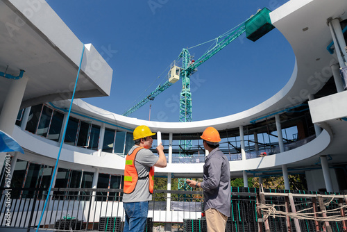 Two young structural engineer and architect dressed in orange work vests standing in front of mechanical crane working on the construction site.