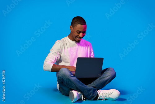 Black man using laptop, sitting on blue in neon light