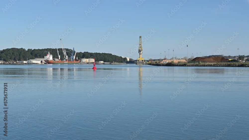Interior Mercantile Port of Bayonne, France, in the foreground the calm water of the river flows, in the background a maroon ship with three white cranes in the middle a red buoy,a large yellow crane Interior Mercantile Port of Bayonne, France, in the foreground the calm water of the river flows, in the background a maroon ship with three white cranes in the middle a red buoy,a large yellow crane