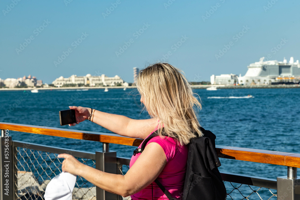 a pice of a blonde tourist girl with a rucksack standing on a Dubai embankment with a stunning view of the sea and surrounding contemporary skyscrapers