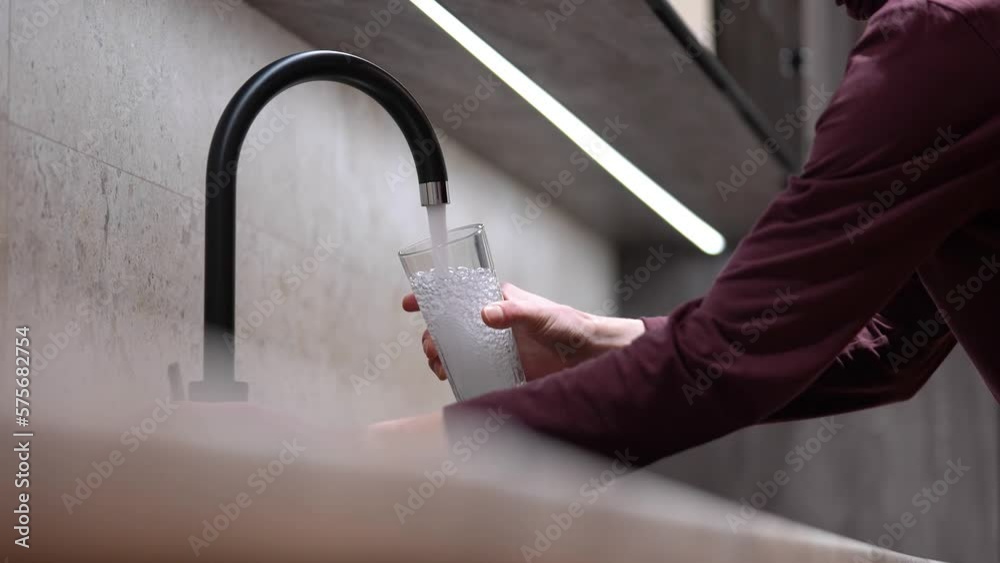 A man is holding a glass, water is flowing from the faucet in the kitchen. Glass with clean water. Concept: Tap water, drinking water ecology, water filters
