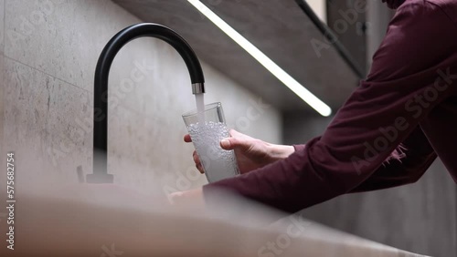 A man is holding a glass, water is flowing from the faucet in the kitchen. Glass with clean water. Concept: Tap water, drinking water ecology, water filters