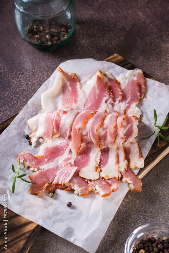 Raw bacon slices on paper on a cutting board on the table. Hearty snack. Vertical view