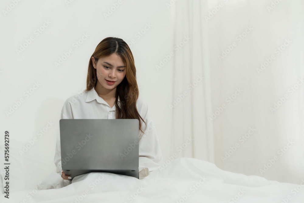 young asian woman using laptop when sitting on a bed. Smart living with communication technology for a better life. girl sitting on at white bedroom space with a clean interior design.