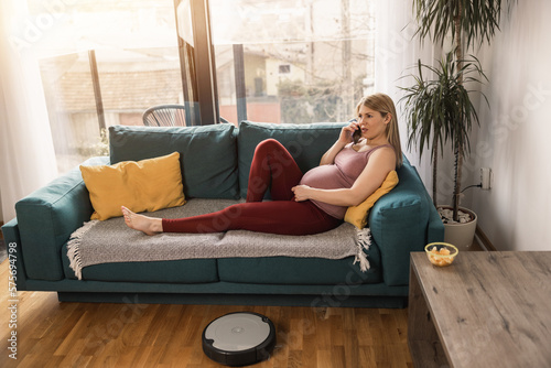 Pregnant Woman Relaxing On Sofa While Robotic Vacuum Cleaner Cleaning At Home