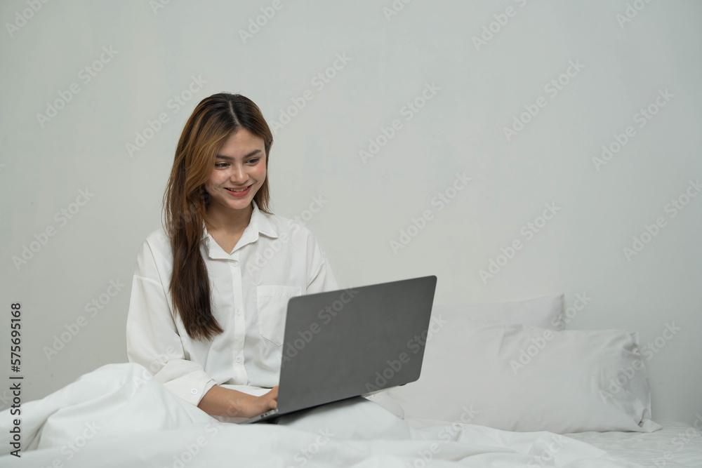 young asian woman using laptop when sitting on a bed. Smart living with communication technology for a better life. girl sitting on at white bedroom space with a clean interior design. young asian woman using laptop when sitting on a bed. Smart living with communication technology for a better life. girl sitting on at white bedroom space with a clean interior design.