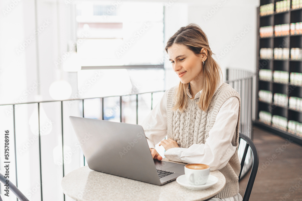 Attractive happy young girl student studying at the college library, sitting at the table with coffee, using laptop computer.Freelancer woman have video call, training coach, teacher lesson. Attractive happy young girl student studying at the college library, sitting at the table with coffee, using laptop computer.Freelancer woman have video call, training coach, teacher lesson.