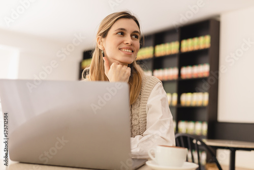 Image of young beautiful joyful woman smiling while working with laptop in office or cafe with coffee on table. Woman wear knitted vest and white shirt, hold her head. Good work day.