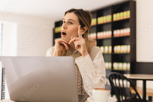 Have idea! Portrait of creative positive attractive young girl freelancer in knitted beige vest and white shirt is sitting in cafe and working on laptop with toothy smile and showing finger up. indoor