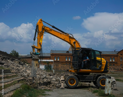 Hydraulic breaker hammer excavator at demolition work. yellow crusher excavator stands sideways on ruins of building against backdrop of rural landscape and blue sky Crushing concrete structures