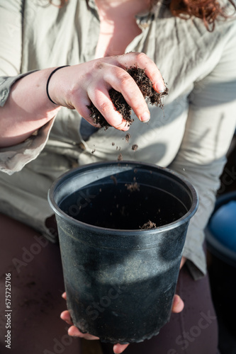 Woman with brown curly hair is putting soil in a black pot with her hands, vertical shot, no visible face