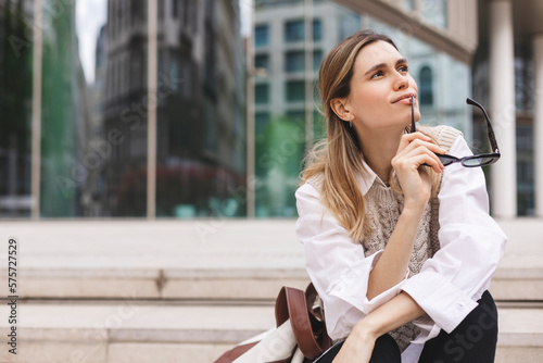 Beautiful smiling fashionable businesswoman sitting on the stairs in front of business center, she look at side, dreaming or thinking, bite shackle of sunglasses. Woman wear knit vest, white shirt.