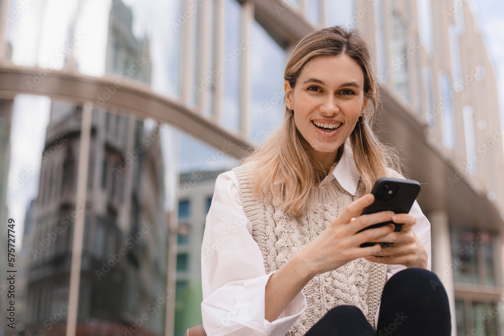 Low angle of glad woman in formal wear sitting in megapolis on street near urban building using smartphone, look happy and smiling. Blonde girl read good news, look at camera.