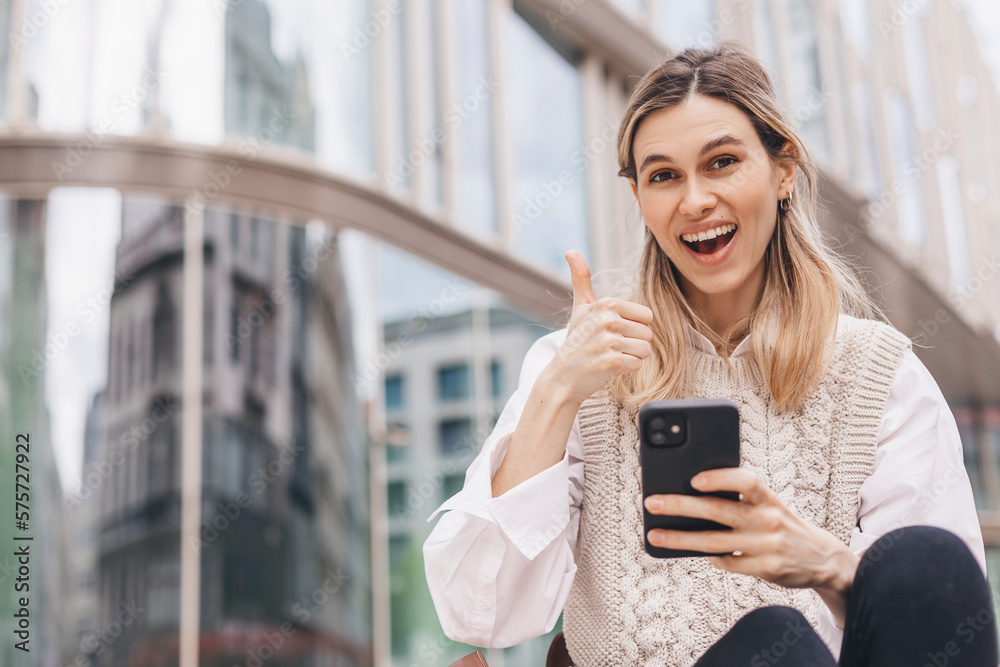 Beautiful smiling fashionable businesswoman sitting on the stairs in front of business center and show thumb up, hold mobile phone, look happy. Amazed woman wear beige knit vest, white shirt and bag. Beautiful smiling fashionable businesswoman sitting on the stairs in front of business center and show thumb up, hold mobile phone, look happy. Amazed woman wear beige knit vest, white shirt and bag.