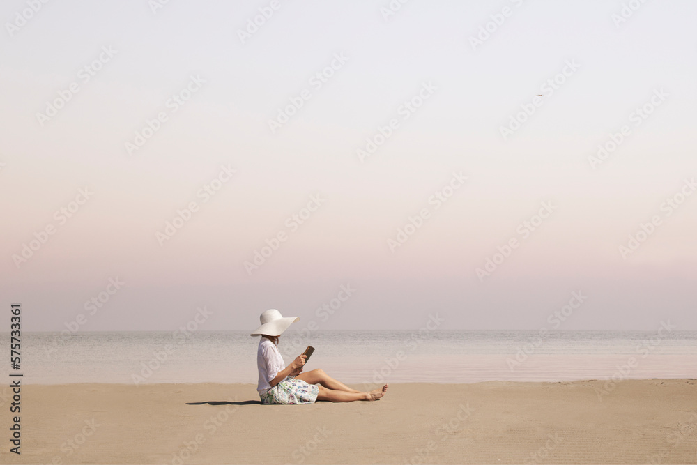 woman with hat reads a book on the beach