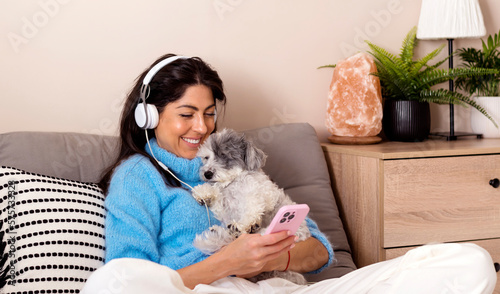 Young woman with headphones sitting at home with her dog listening ebook with her mobile phone 