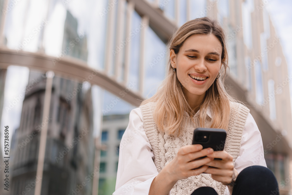 Low angle of glad woman in formal wear sitting in megapolis on street near urban building using smartphone, look happy and smiling. Blonde girl read good news.